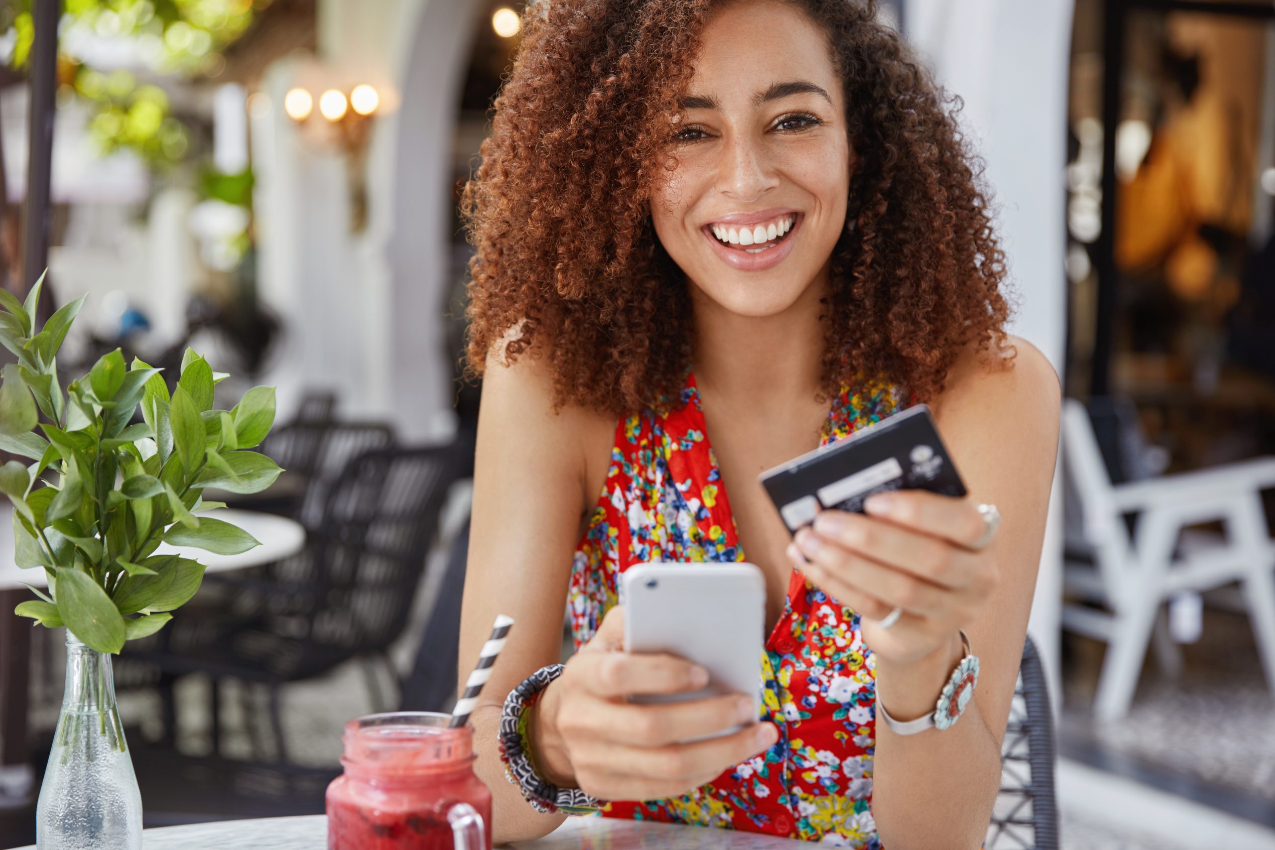 Internet banking and ecommerce concept. Happy young smiling female with Afro hairstyle, uses modern cell phone and credit card for online shopping, enjoys fresh fruit smoothie in terrace bar.