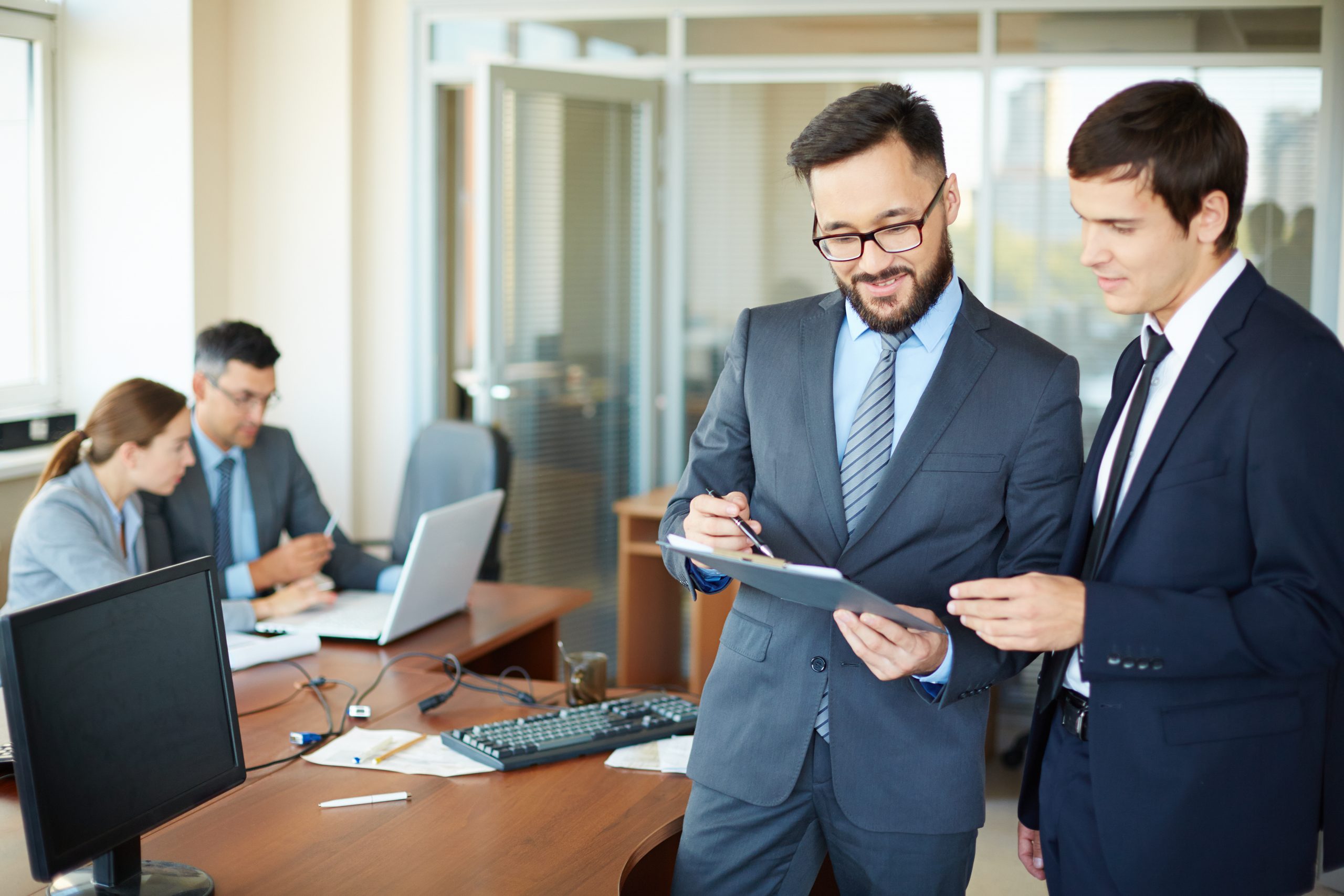 Confident businessman with clipboard consulting his partner on background of their colleagues in office